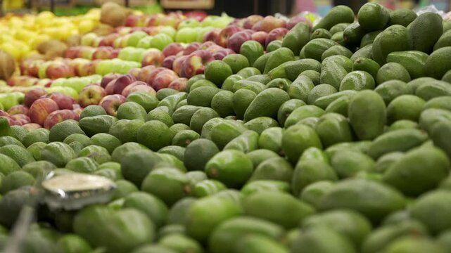 Mound of green avocados fills the foreground with rows of apples and lemons behind on a supermarket produce aisle.
