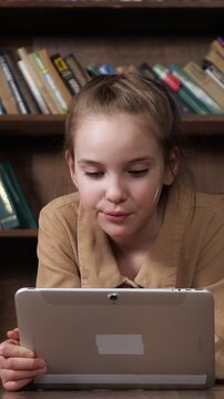 Focused little girl using a modern digital tablet for online entertainment or remote learning, lying on the floor in a library with a large bookshelf full of classic books in the background