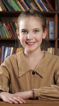 Adorable schoolgirl with blue eyes sitting at her desk in a classroom or library. Cute little pupil looking at the camera and smiling, ready for learning, with bookshelves in the background