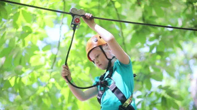 Young girl crossing an obstacle on a ropes course in an adventure park, wearing a helmet and safety harness among trees