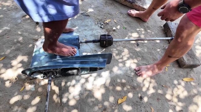 close-up high angle shot of two workers barefoot performing a collaborative field repair on an outboard motor lower unit using a slide hammer on a beach.