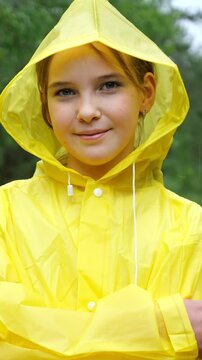 Portrait of a happy young girl with blue eyes wearing a bright yellow hooded raincoat, smiling confidently at the camera while standing outdoors in a park or forest during wet weather