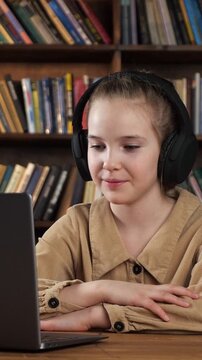 Smiling schoolgirl wearing headphones using a laptop for an online lesson in a library, actively participating in a video call with her teacher for a remote education session