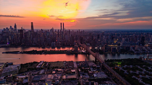 New York City skyline at sunset, view of Manhattan and Queensboro Bridge. Aerial view of the East River and skyscrapers under an orange sky.