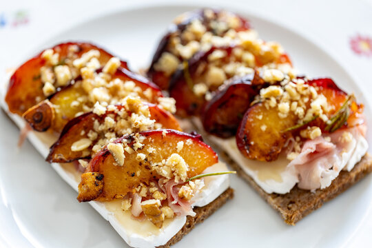 Snack with a slice of bread, fried peach, camembert, prosciutto and crushed walnut on a white plate with flowers