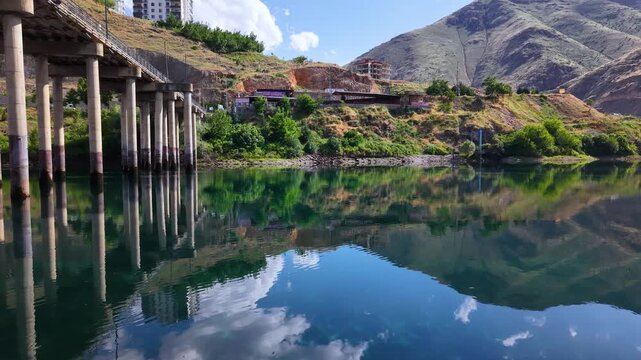 Smooth lateral aerial movement passes beside massive bridge piers above a calm lake, where crystal-clear reflections and scattered clouds create a stunning mirrored waterscape.