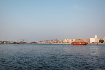 Fototapeta premium A scenic view of a traditional wooden boat sailing on the calm waters of Dubai Creek with city buildings and a clear blue sky in the background in Dubai, United Arab Emirates. 