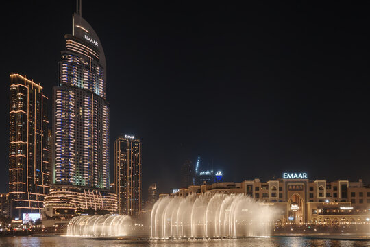 A night view of the illuminated fountains at Dubai Fountain Lake with the EMAAR building and skyscrapers in the background in Dubai, United Arab Emirates.