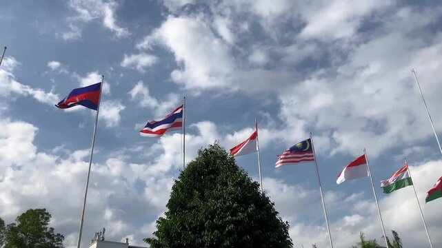 Cinematic slow motion of Asian and Australian national flags including Thailand, Malaysia, and Indonesia waving on flagpoles against a partly cloudy blue sky.
