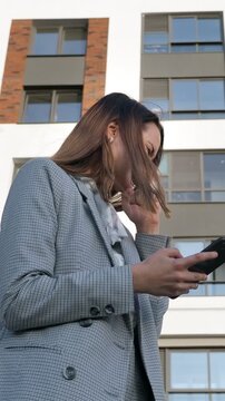 Successful real estate agent celebrating a victory after checking her smartphone, exulting with a fist pump in front of a modern residential apartment building complex