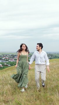 Cheerful young couple in love holding hands and running through a picturesque grassy field on a hilltop, smiling and looking at each other with a scenic countryside village below