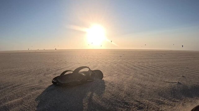 Windswept flip-flops on the beach during a golden hour sandstorm with distant kiteboarding action