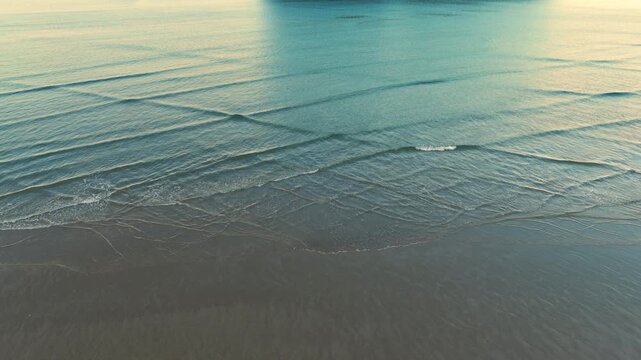 A beautiful aerial drone view of soft ocean waves rolling onto the sandy shoreline at Whangamata, Coromandel Peninsula, New Zealand, during golden hour.