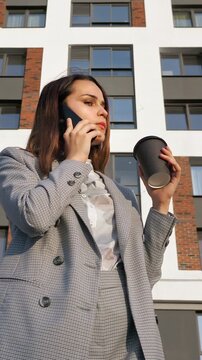 Young successful businesswoman or realtor talking on a smartphone and drinking coffee, standing against a modern residential building, confidently discussing business and smiling