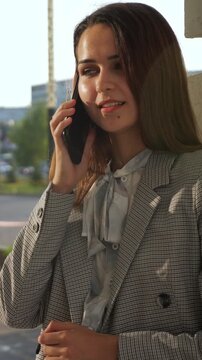 Beautiful smiling businesswoman in a stylish blazer having an engaging conversation on her mobile phone, her cheerful expression highlighted by the warm sunlight outdoors in a city setting