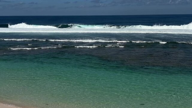 A serene beach scene with clear turquoise water and gentle waves. The horizon features a blue sky with scattered clouds.
