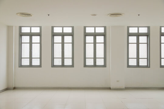 A clean, minimalist interior of an empty white room featuring a row of classic grey-framed windows, white tiled floor, and ventilation vents on the ceiling, architectural background concept.