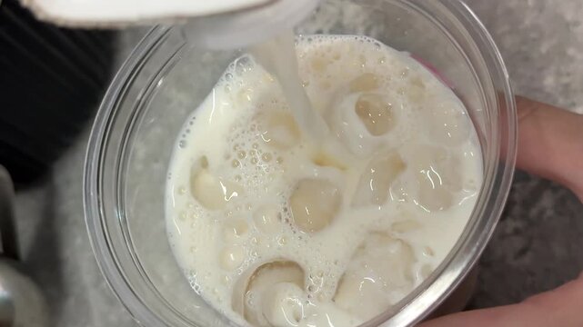 Pouring milk into plastic cup filled with ice, refreshing cold dairy drink preparation for iced latte or milk beverage close up