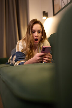 Woman lying on green sofa looking surprised at phone screen