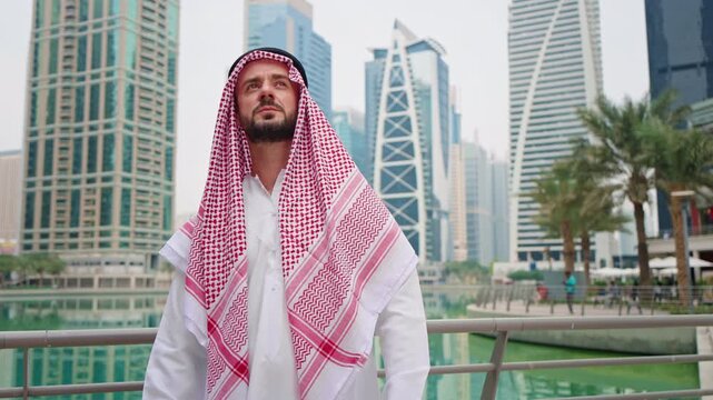 Arabic businessman in a traditional robe and a headscarf, looking at modern buildings while standing in a business Gulf city center.