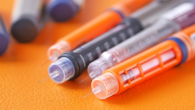 Close up view of insulin pens with needles on an orange background, several other pens are blurred in the background, a focus on medical devices for diabetes management