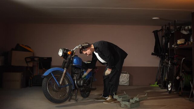 Young man wearing work clothes and gloves checking and repairing a classic vintage blue motorcycle in a home garage, focusing on engine maintenance