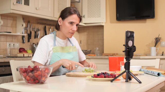 Woman preparing fresh fruit and berries in her kitchen, recording the process with a smartphone on a tripod for a live streaming or online cooking class