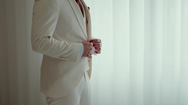 Man in cream suit holding pocket watch chain standing near window