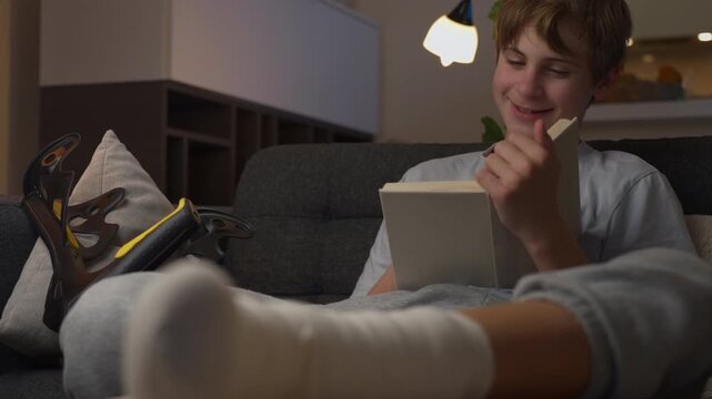 Teenage boy recovering from leg injury, relax on sofa and read book with bandaged leg elevated. Home recovery, rehabilitation, rest and healing process concept.