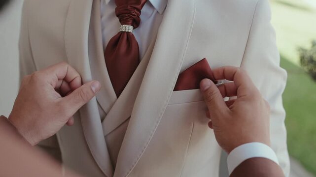 Hands placing rust colored pocket square into cream suit jacket pocket
