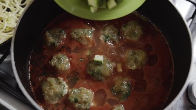 Top view of adding diced zucchini into simmering meatball sauce while preparing homemade meal on kitchen stove in home kitchen.