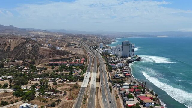 Aerial flyover above Transpeninsular Federal Highway 1 approaching Club Marena Real Estate on the Rosarito coastline, Baja California, Mexico with Pacific Ocean surf