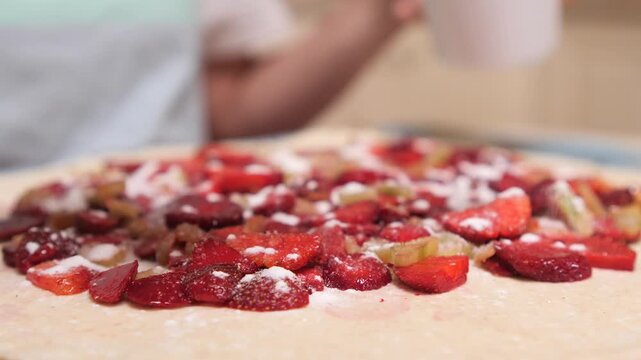 Person preparing a sweet fruit pie, sprinkling sugar from a white cup onto chopped strawberries and rhubarb laid out on a sheet of pastry dough, ready for baking