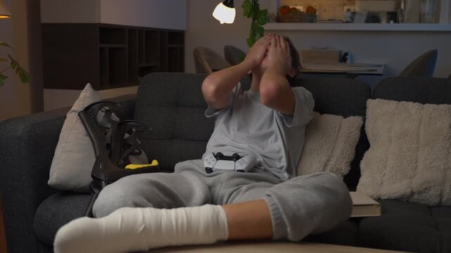 Frustrated teenage boy with a broken leg in a cast reacting to losing a video game while sitting on a sofa at home. Gaming, disappointment, teenage emotions and injury recovery concept.