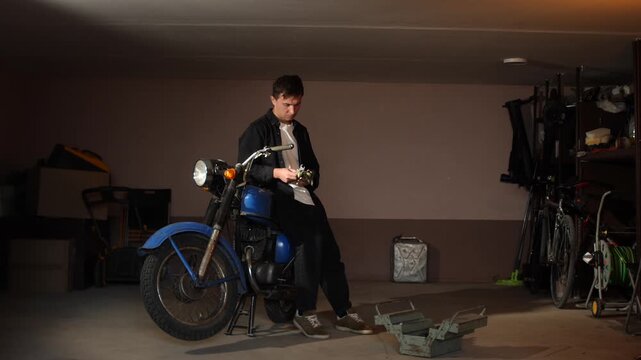 Man feeling weary, wiping his forehead while leaning on a vintage 1970s motorcycle, taking a short break from restoration work in a dimly lit garage setting