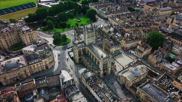 High aerial drone footage over Bath, England, showing Bath Abbey's full length and tower from above, the adjoining Abbey Churchyard square with visitors, the Roman Baths' domed roof nearby