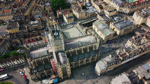 Top-down aerial drone footage over Bath, England, showing Bath Abbey's rooftop and Gothic tower from directly above, the adjoining church square with visitors, and the domed Roman Baths complex beside