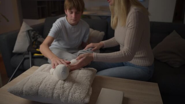 Mother providing supportive care to teenage son with a broken leg in a plaster cast at home. Injury recovery, patient comfort, rehabilitation and home healthcare concept.