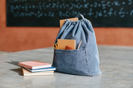 Denim drawstring backpack with school supplies and notebooks on desk in classroom for education background and product display