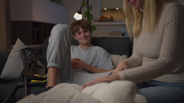Loving mother carefully applying a bandage to her teenage son's injured leg as he rests on the couch with a foot brace, representing family care, support, and recovery at home