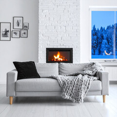 Obraz premium A cozy living room interior featuring a modern grey sofa, warm fireplace, knit blanket, and snowy winter forest window view, evoking warm comfort and serene peace. There are no people.