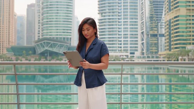Portrait of a confident real estate agent Asian businesswoman using a tablet to work on business, with modern skyscrapers of a Gulf city