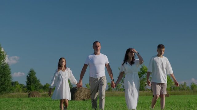 A full-length view of a happy family - mother, father, son and daughter walking forward hand-in-hand through a green field on sunny day. Peaceful moment of family togetherness and rural lifestyle.