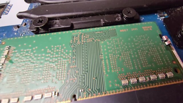 Close-up of a laptop RAM memory card circuit board during repair, showing gold-plated connector pins, soldered components, and internal computer hardware