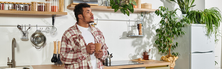 Man stands in a stylish kitchen, holding a cup and gazing thoughtfully while surrounded by plants. © LIGHTFIELD STUDIOS