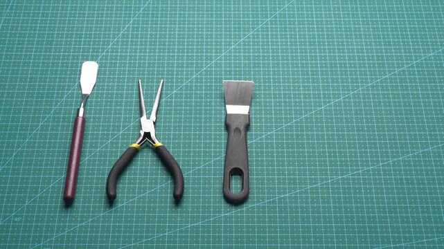 Four precision hand tools&mdash;spatula, needle-nose pliers, putty scraper, and wire cutters&mdash;arranged on green self-healing cutting mat for DIY electronics repair or model crafting