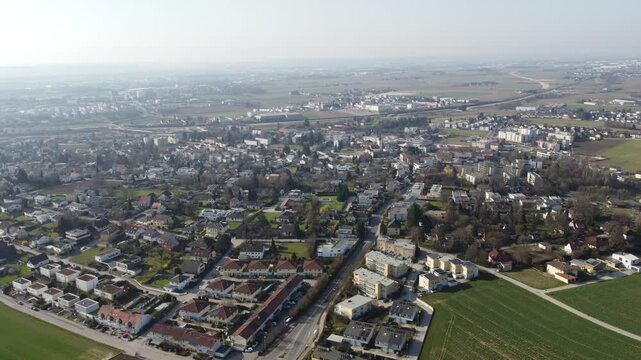 Aerial drone view of Leonding, Austria, showing a residential town with houses, apartment buildings, solar-paneled rooftops, and streets bordered by farmland under a hazy sky.