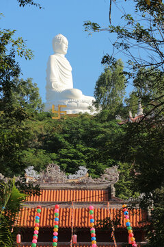 Giant White Buddha statue sitting on a hilltop above the traditional tiled roof of Long Son Pagoda temple.