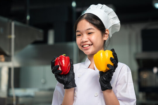 Junior Chef Holding Fresh Red and Yellow Bell Peppers While Smiling in a White Culinary Uniform Inside a Modern Commercial Training Kitchen for Culinary Education