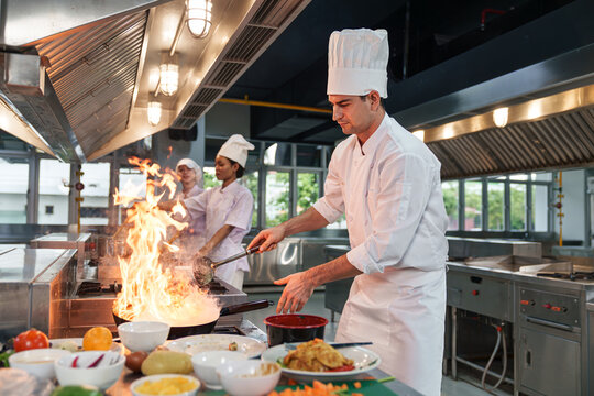 Restaurant Chef Performing Flambe Cooking with Flames Rising from a Pan While Two Black Chefs Work Behind Him in a Modern Commercial Kitchen